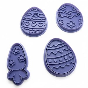 May include: Four purple Easter-themed cookie cutters. Designs include an egg with hearts, an egg with a chick hatching, a carrot with flowers, and an egg with zigzags and dots. Perfect for spring baking.
