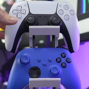 Op de afbeelding: Een wit en zwart PlayStation 5-controller is gestapeld boven een blauwe Xbox-controller op een witte standaard. De controllers hebben verschillende knoppen en joysticks. De achtergrond is wazig, met een paarse tint.