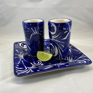 Puede incluir: Dos vasos de chupito de cerámica azul y blanca con una bandeja cuadrada de cerámica azul y blanca y una rodaja de lima.