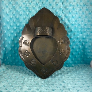 Op de afbeelding: Een metalen hartvormige wanddecoratie met een gegolfde rand en bloemmotieven. Het hart heeft een kleine, rechthoekige opening aan de bovenkant, die gebruikt kan worden voor een kaars of een ander klein voorwerp.