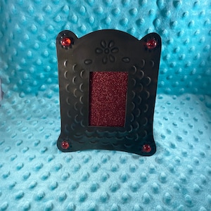 May include: Black metal picture frame with a red glitter background and red glass accents. The frame has a scalloped design and a flower cutout at the top.