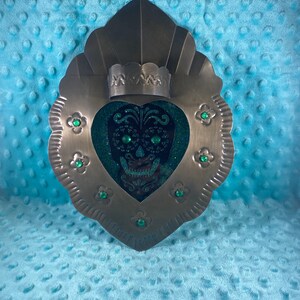 May include: A heart-shaped metal wall decor with a green glitter skull design and green gem accents. The decor has a crown-like top and a scalloped edge.