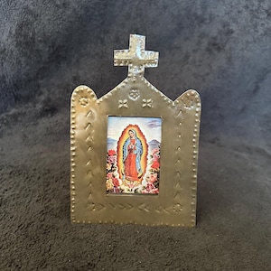 May include: A small, metallic religious icon featuring an image of the Virgin Mary. The frame is a light gold color with a cross at the top and decorative edging. The image within the frame depicts the Virgin Mary surrounded by roses.