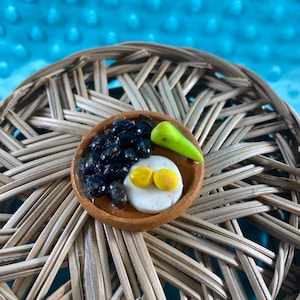Puede incluir: Un pequeño plato de arcilla con frijoles negros, un disco blanco y dos círculos amarillos, coronado con un triángulo verde. El plato está sobre una superficie de mimbre tejido.