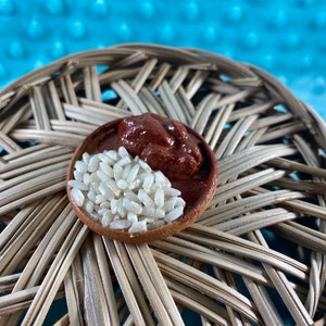 Puede incluir: Un pequeño cuenco marrón lleno de arroz blanco y una cucharada de salsa marrón. El cuenco está sobre una superficie de mimbre tejido.