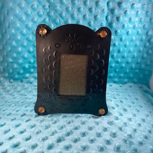 May include: Black metal picture frame with a decorative design and gold accents. The frame has a textured surface and a cutout for a photo.