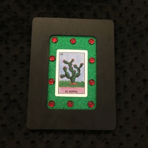 May include: A black framed card with a green background and red glitter. The card features a traditional Mexican Loteria card with the number 39 and the image of a cactus. The card reads "El Nopal".
