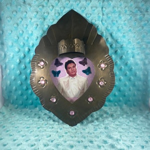 May include: A heart-shaped metal frame with a photo of a man in a white tuxedo. The frame is decorated with pink rhinestones and butterflies.
