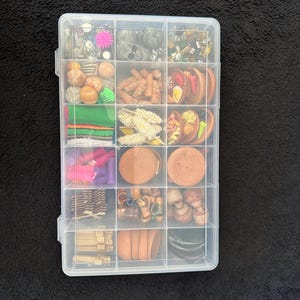 Może przedstawiać: Przezroczyste plastikowe pudełko z miniaturowymi produktami spożywczymi i materiałami rzemieślniczymi. Komory zawierają małe, szczegółowe repliki żywności, takiej jak sushi, jajka i ciasta, wraz z innymi elementami dekoracyjnymi. Pudełko znajduje się na czarnej powierzchni.