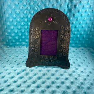May include: A silver metal picture frame with a decorative floral design and a pink rhinestone accent. The frame has a purple glitter background.