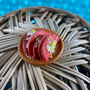 Puede incluir: Un plato de arcilla en miniatura con tres tacos rojos cubiertos con ingredientes blancos y verdes.