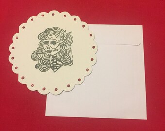 May include: A white card with a scalloped edge and red glitter dots. The card features a black and white illustration of a woman with a skull face and flowers in her hair.