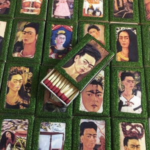 May include: A collection of matchboxes featuring various images of Frida Kahlo, a famous Mexican painter. The matchboxes are decorated with green glitter and have a green border. One matchbox is open, revealing the matches inside.