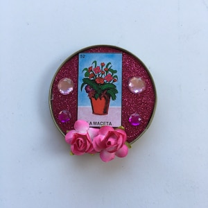 May include: A pink and gold round card with a flower pot and flowers on it. The card has the number 52 and the words "La Maceta" on it. The card is decorated with pink glitter and pink rhinestones.