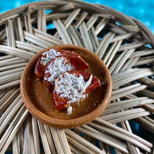 Puede incluir: Un pequeño plato de terracota con tres rodajas de fruta roja cubiertas de copos de coco blanco.