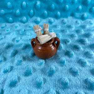 Puede incluir: Una pequeña olla de cerámica marrón con dos manojos de hierbas secas atadas con hilo blanco. La olla tiene dos asas pequeñas y descansa sobre una superficie azul claro texturizada.