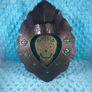 May include: A metal heart-shaped wall decor with a gold sugar skull cutout and green glitter. The skull has blue eyes and is surrounded by green gems.