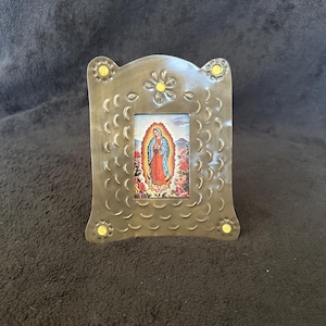 Könnte beinhalten: Kleiner, dekorativer Bilderrahmen aus Metall mit einem religiösen Bild. Der Rahmen ist silberfarben mit einem strukturierten Muster und gelben Akzenten. Das Bild zeigt eine religiöse Figur in einer farbenfrohen Umgebung.