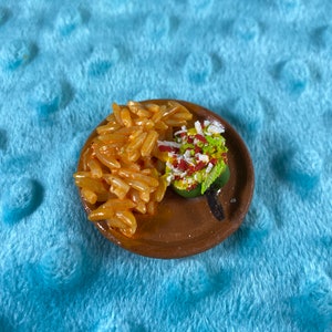 Puede incluir: Un plato de arcilla en miniatura con una pequeña porción de arroz rojo y un chile verde cubierto con queso blanco y salsa roja.