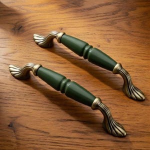 Puede incluir: Dos tiradores de armario con una sección central cilíndrica de color verde oscuro liso y extremos de metal con acabado en latón antiguo. Los extremos presentan un diseño acampanado en forma de abanico con crestas detalladas.