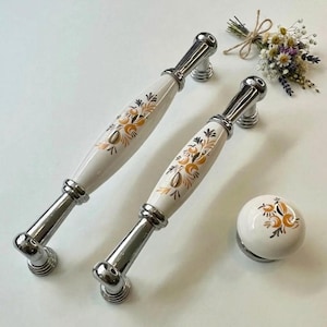 Op de afbeelding: Set van twee witte en goudkleurige kastgrepen met bloemmotief en een bijpassende knop. De grepen hebben een chromen afwerking op de basis en de uiteinden van de handgrepen. Het bloemmotief is in goud- en bruintinten.