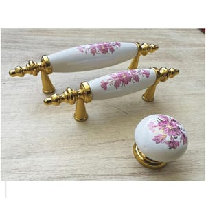 Puede incluir: Juego de dos tiradores de armario blancos y dorados y un pomo a juego. Los tiradores tienen un diseño curvo con un centro de porcelana blanca con un estampado floral rosa. Los herrajes son dorados.