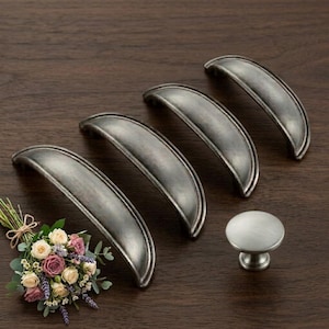 May include: Four antique-style cabinet pulls and one knob in a distressed silver finish. The pulls are curved with a raised edge, and the knob is round. A small bouquet of flowers is in the lower left corner, set against a dark wood surface.