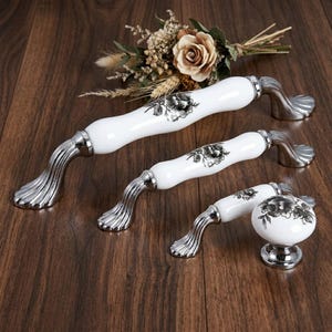 Può includere: Set di tre maniglie per mobili in ceramica bianca con basi in metallo color argento. Le maniglie presentano un design floreale nero. La ferramenta per mobili è disposta su una superficie in legno scuro, con un bouquet di fiori secchi sullo sfondo.