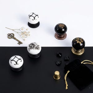 Puede incluir: Un surtido de pomos decorativos para armarios con diseños florales y geométricos en blanco y negro. Otros elementos incluyen una llave antigua, piedras negras, un dedal y una pequeña bolsa de terciopelo negro con cordón dorado.