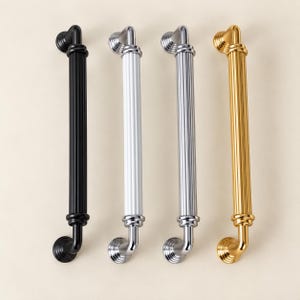 Peut inclure: Quatre poignées de placard verticales disposées côte à côte. Chaque poignée présente un centre cylindrique cannelé avec des embouts arrondis. Les finitions de gauche à droite sont noir mat, blanc brillant, chrome poli et métal doré poli.