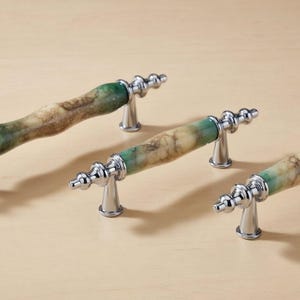 Op de afbeelding: Drie kastgrepen met een natuurstenen uitstraling. De handgrepen zijn een mix van groen, bruin en beige, met verchroomde metalen accenten. De grepen zijn ontworpen met een cilindrische vorm en zijn gemonteerd op kleine, decoratieve voetjes.