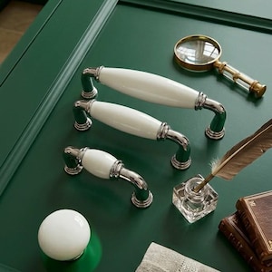Peut inclure: Trois poignées de meubles blanches et argentées, une loupe, un encrier et une plume sur une surface vert foncé. Les poignées ont un design classique avec une poignée en céramique blanche et des accents argentés.