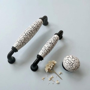 May include: Two cabinet handles and a knob. The handles have a white, crackled ceramic design with black accents and a black metal base. The knob has a matching white crackled ceramic design. A few dried flowers are in the image.