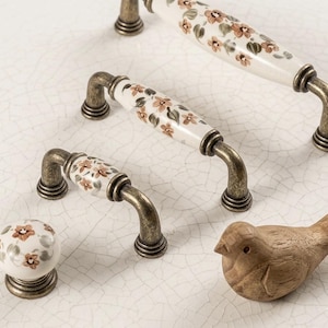 May include: A selection of antique-style cabinet handles and a knob. The handles feature a cream ceramic centre with a floral pattern in brown and green, and bronze-coloured metal fixtures. A small wooden bird is also present.