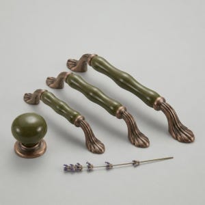Puede incluir: Un conjunto de herrajes para armarios en verde oliva y bronce, que incluye tres tiradores y un pomo redondo. Los tiradores tienen un centro curvo, similar a la cerámica, y extremos de bronce ornamentados. También hay una ramita de lavanda.