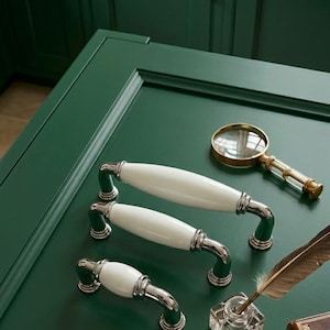 May include: A collection of cabinet hardware and accessories on a dark green surface. Includes white and chrome cabinet pulls, a magnifying glass, an inkwell, a feather quill, and a stack of books. The cabinet pulls have a white ceramic center.