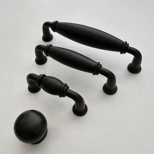 Op de afbeelding: Een set zwarte kastbeslagen, waaronder twee handgrepen, een kleinere handgreep en een ronde knop. De handgrepen hebben een gebogen ontwerp met decoratieve details. De hardware is gemaakt van matzwart materiaal.