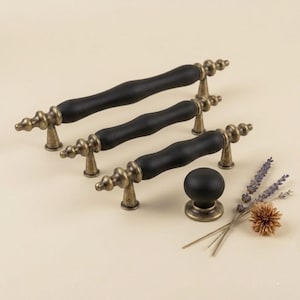 May include: A set of black and antique brass cabinet hardware. The set includes three different-sized handles and a round knob. The handles have a black, cylindrical center and ornate brass ends. The knob is a solid black sphere with a brass base.