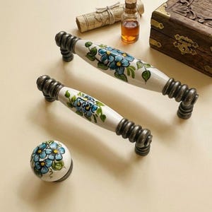 May include: Two white ceramic cabinet pulls and a knob with a floral design in blue and green. The pulls have a metal base and are oval-shaped. The knob is round. The cabinet hardware is set against a neutral background.