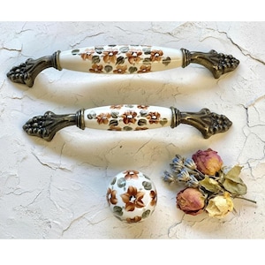 Può includere: Due maniglie per mobili bianche e marroni con motivi floreali e estremità a grappolo d'uva color bronzo. Sono presenti anche un pomello rotondo abbinato e fiori secchi. Le maniglie misurano circa 15 cm di lunghezza.
