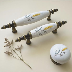 May include: Set of two white ceramic cabinet handles and a matching knob. Each handle features a wheat design in yellow and gray, with antique bronze-colored metal hardware. The knob has the same wheat design.