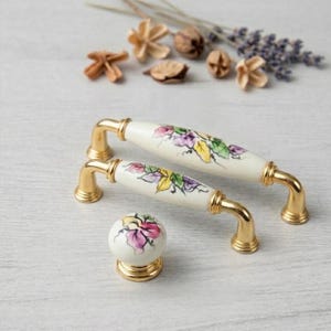 以下が含まれることがあります： 3つの装飾的なキャビネット金具のセット。 金色のアクセントと花の模様が付いた2つの長い白い磁器製のハンドルと、お揃いの丸いノブ。 花柄のデザインは、紫、黄色、緑の葉と花が特徴です。