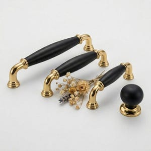May include: Cabinet hardware set with three black and gold handles and a knob. The handles have a black, cylindrical centre and gold-coloured, curved ends. A small bouquet of dried flowers is placed near the hardware.
