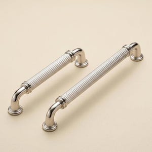 Könnte beinhalten: Zwei Schrankgriffe aus gebürstetem Nickel. Jeder Griff hat einen geriffelten Mittelteil und gebogene Enden. Die Griffe sind für eine einfache Installation konzipiert und verleihen jedem Schrank oder jeder Schublade einen Hauch von Eleganz.