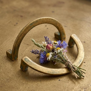 May include: Two antique brass drawer pulls with a curved design, resting on a textured surface. A small bouquet of wildflowers, including daisies, lavender, and dried roses, is nestled between the pulls, tied with twine.