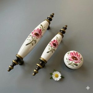 May include: A set of white ceramic cabinet pulls and a knob, each adorned with a pink floral design. The hardware features brass-toned accents and a decorative finial. A small daisy is placed near the hardware.