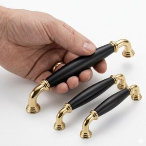 May include: Black and gold cabinet pulls. The pulls have a black center and gold-colored curved ends. One pull is held in a hand, with two others below it. The pulls are likely for kitchen or bathroom cabinets.