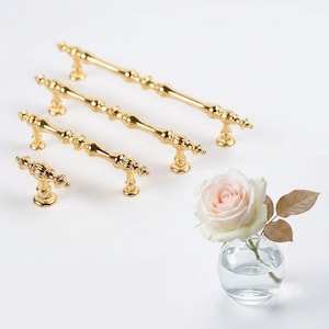 Könnte beinhalten: Eine Sammlung goldfarbener Schrankgriffe und -knöpfe in verschiedenen Größen. Die Hardware hat ein verziertes, Vintage-Design. Eine kleine Glasvase mit einer rosa Rose und braunen Blättern befindet sich im Hintergrund.