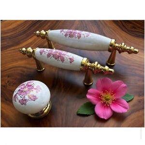 May include: White and gold cabinet hardware set featuring two pull handles and a round knob. The handles and knob have a floral design in shades of pink and purple. The hardware is set on a wooden surface with a pink flower.
