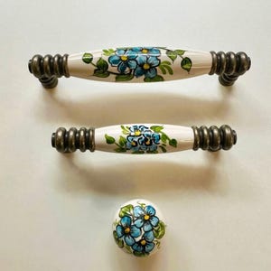 Puede incluir: Tres piezas de herrajes: dos tiradores de armario y un pomo. Los tiradores y el pomo son blancos con un diseño floral de flores azules y hojas verdes. Los herrajes tienen un acabado bronce.
